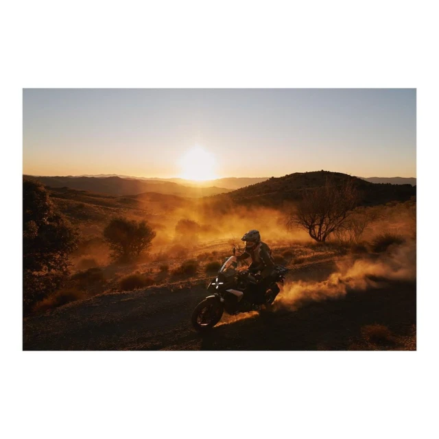 A motorcycle rider navigates a dirt road during golden hour, with dust kicking up behind the bike and a scenic landscape backdrop.