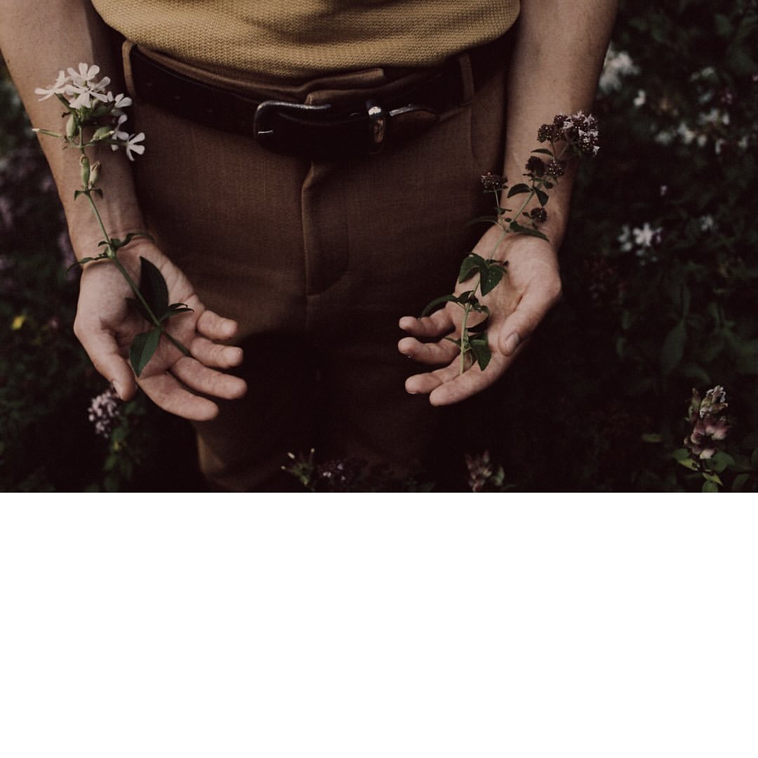 Close-up of a person holding flowers with outstretched hands in a garden with brown pants, a tan top, and a dark belt.
