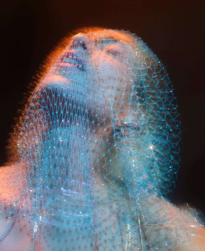 Abstract portrait of a woman's face covered in a sparkling aqua-blue mesh fabric, with soft orange lighting accents against a dark background.