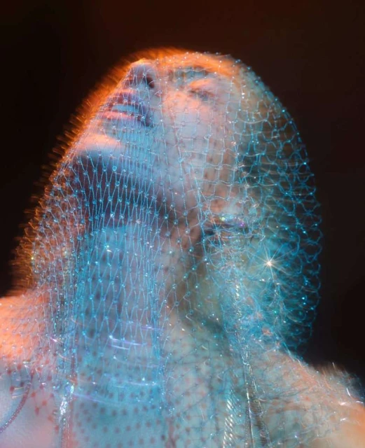 Abstract portrait of a woman's face covered in a sparkling aqua-blue mesh fabric, with soft orange lighting accents against a dark background.
