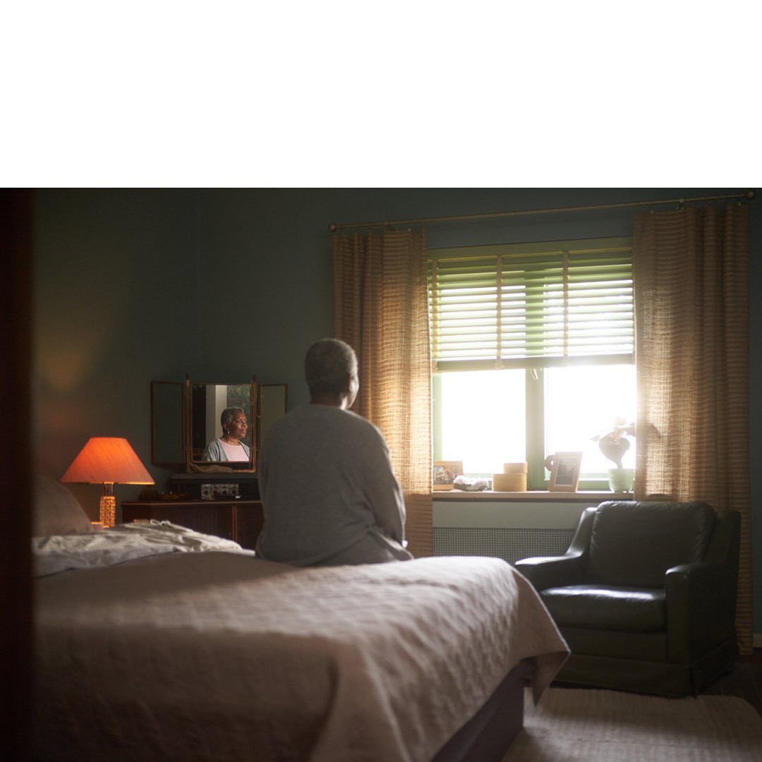 Elderly person sits on a bed in a bedroom, facing a window with natural light. An orange lamp and armchair are visible.