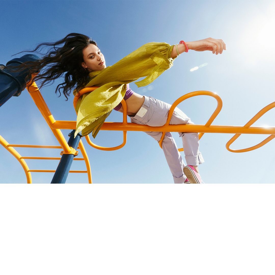 Young woman in a chartreuse top and lavender pants poses on a playground merry-go-round with a blue sky in the background.