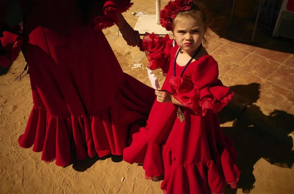 Young girl in red flamenco dress and flower headpiece, holding hands with an adult in red flamenco dress, in outdoor setting.