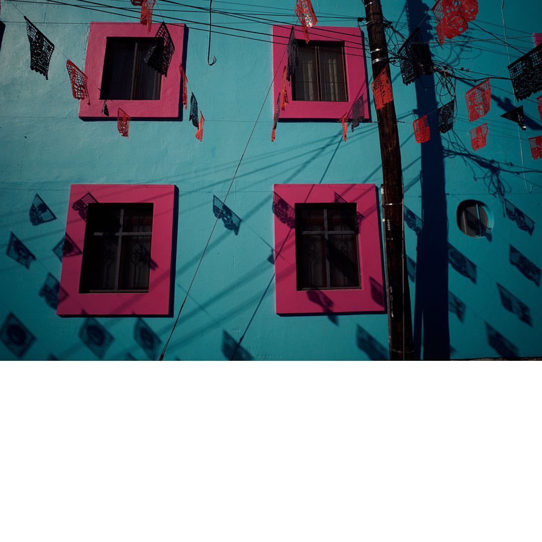 Bright blue building facade with pink window frames and colorful hanging paper decorations, casting shadows in the daylight.