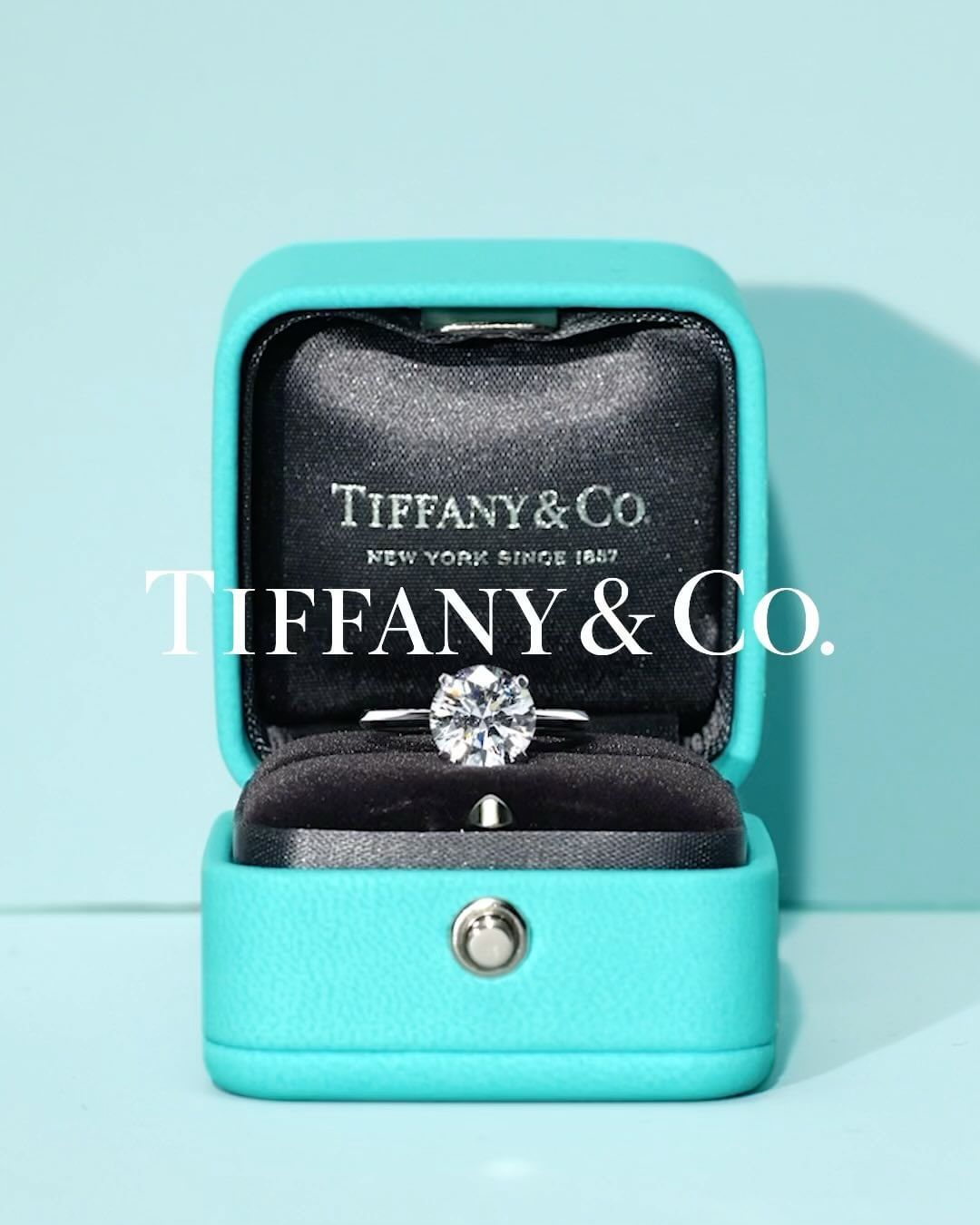 Close-up of a Tiffany & Co. engagement ring with a round brilliant diamond in a signature turquoise blue box on a light blue background.