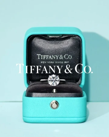 Close-up of a Tiffany & Co. engagement ring with a round brilliant diamond in a signature turquoise blue box on a light blue background.