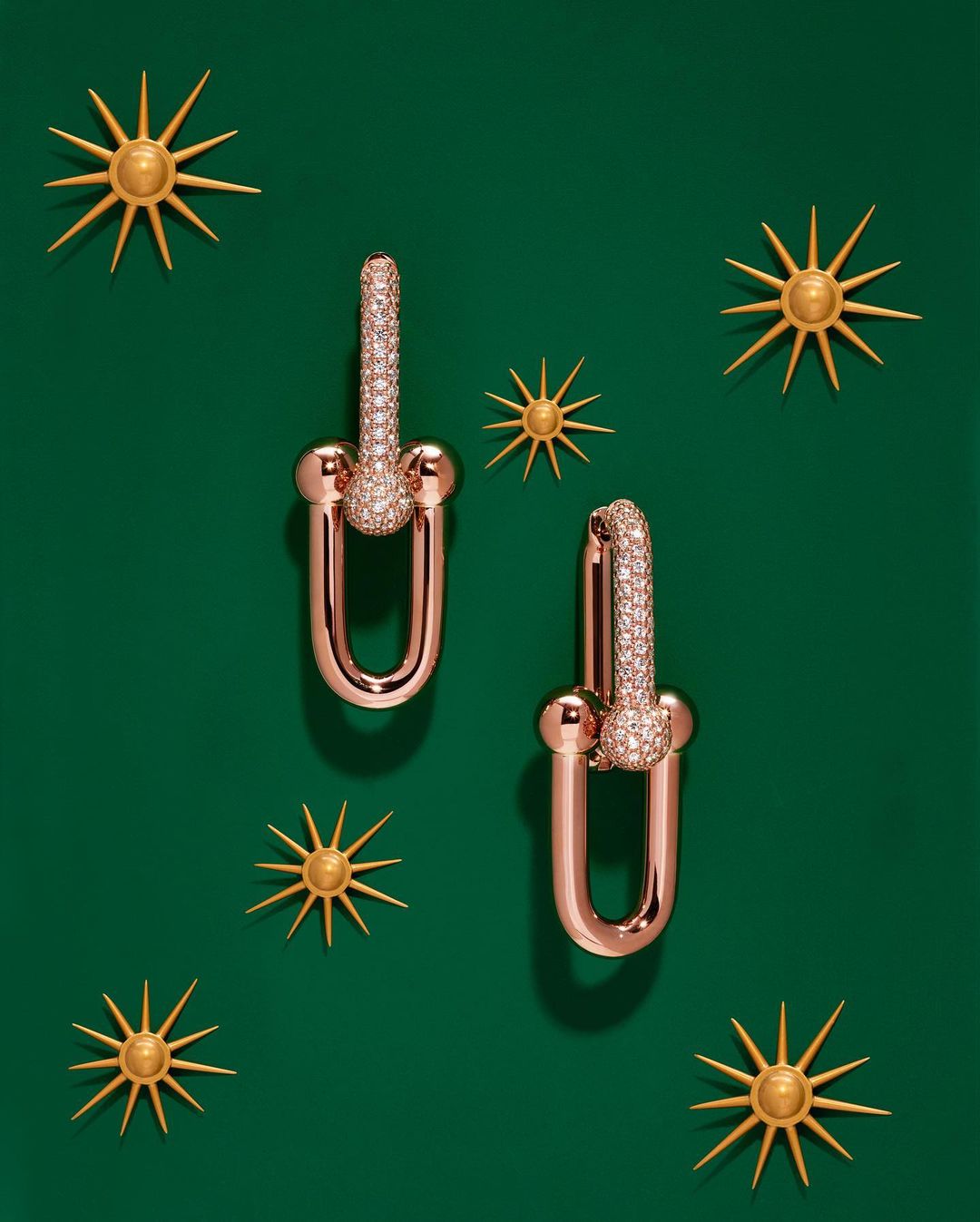 Pair of rose gold Tiffany HardWear link earrings adorned with pavé diamonds, set against a dark green backdrop with sunburst accents.