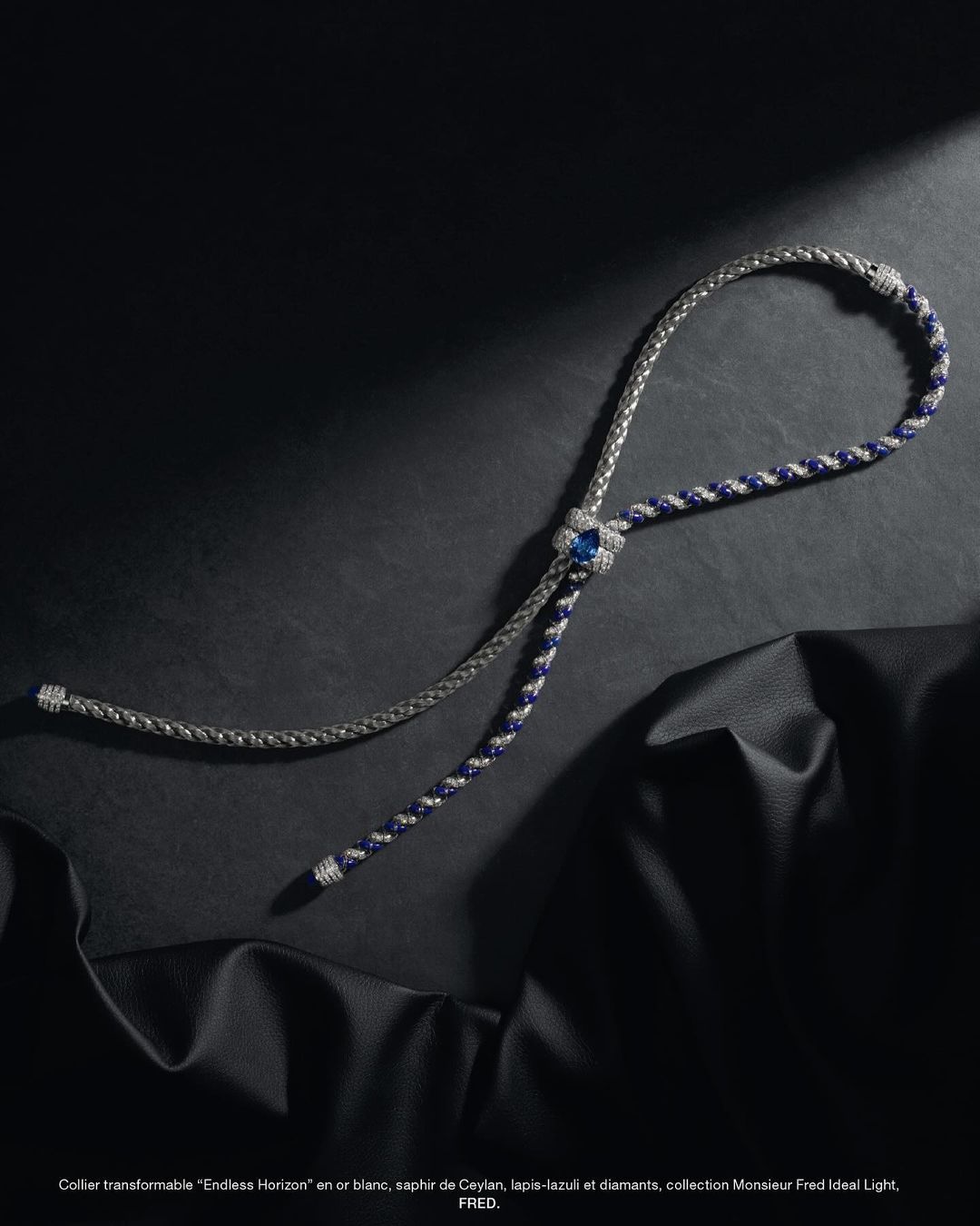 Elegant bolo tie style necklace featuring white gold, sapphires, lapis lazuli, and diamonds, set against a draped black leather surface.