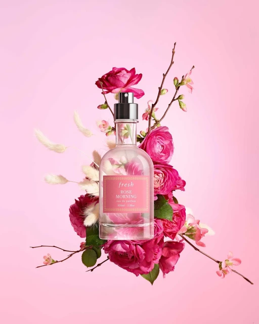 Elegant perfume bottle surrounded by vibrant pink roses and delicate floral branches on a soft pink background, product photography.