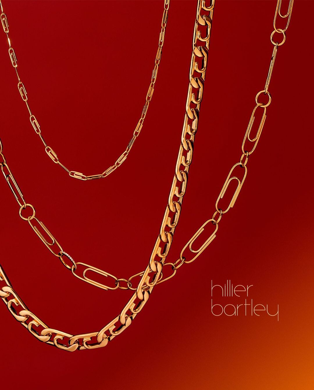 Still life shot of layered gold necklaces and paper clip chains against a red backdrop, illuminated by a subtle gradient lighting effect.