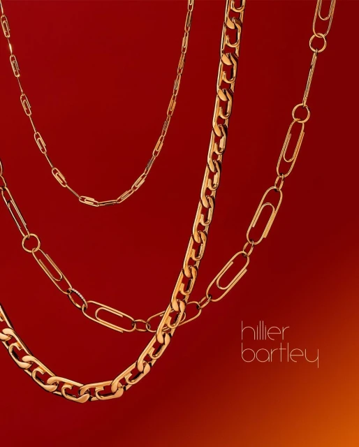 Still life shot of layered gold necklaces and paper clip chains against a red backdrop, illuminated by a subtle gradient lighting effect.