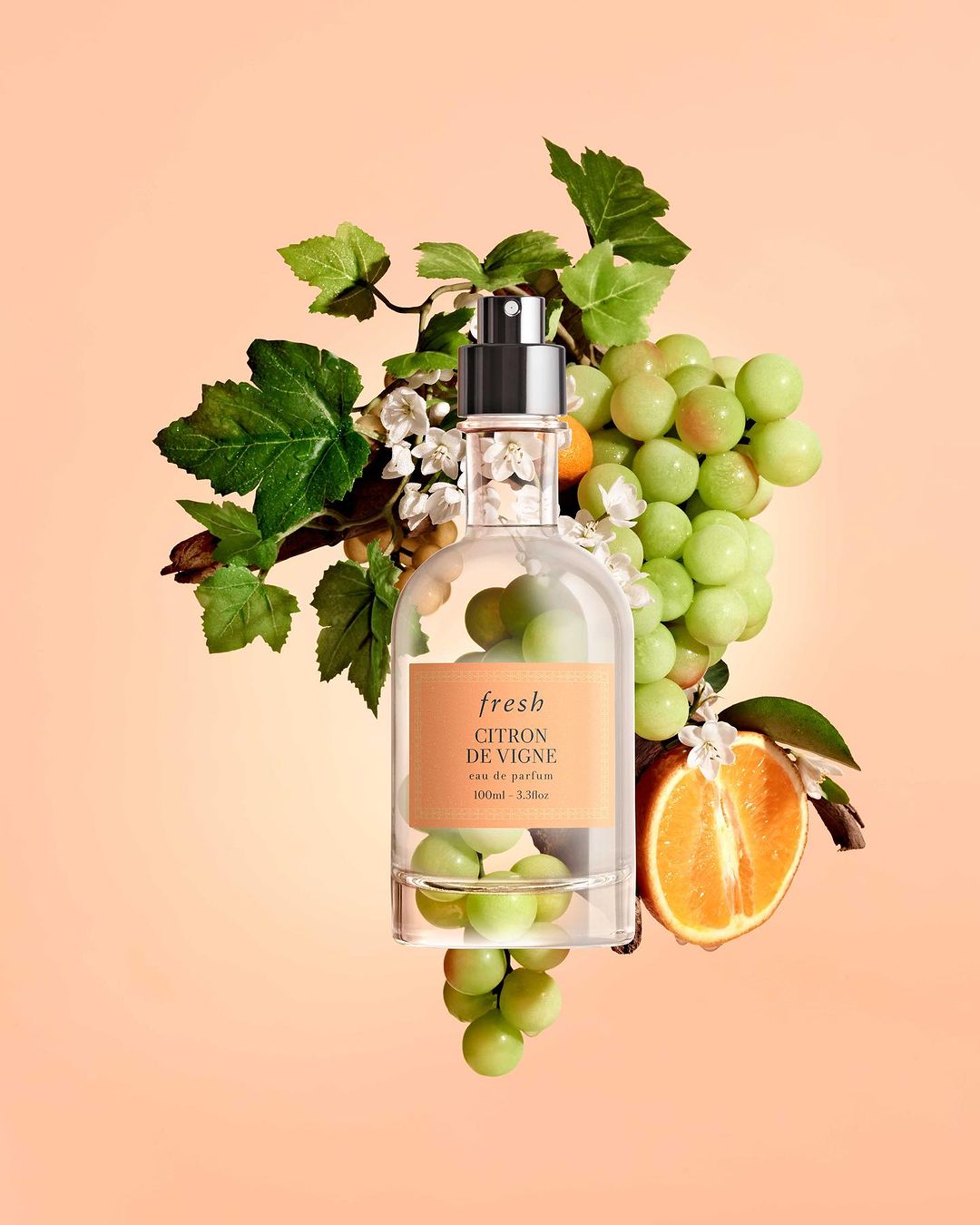 Studio shot of Fresh Citron de Vigne perfume bottle surrounded by grapes, grape leaves, white flowers, and orange half against peach backdrop.