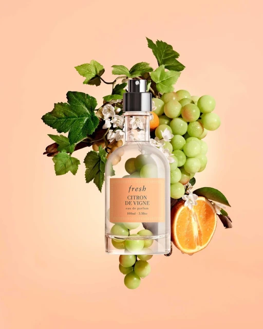 Studio shot of Fresh Citron de Vigne perfume bottle surrounded by grapes, grape leaves, white flowers, and orange half against peach backdrop.
