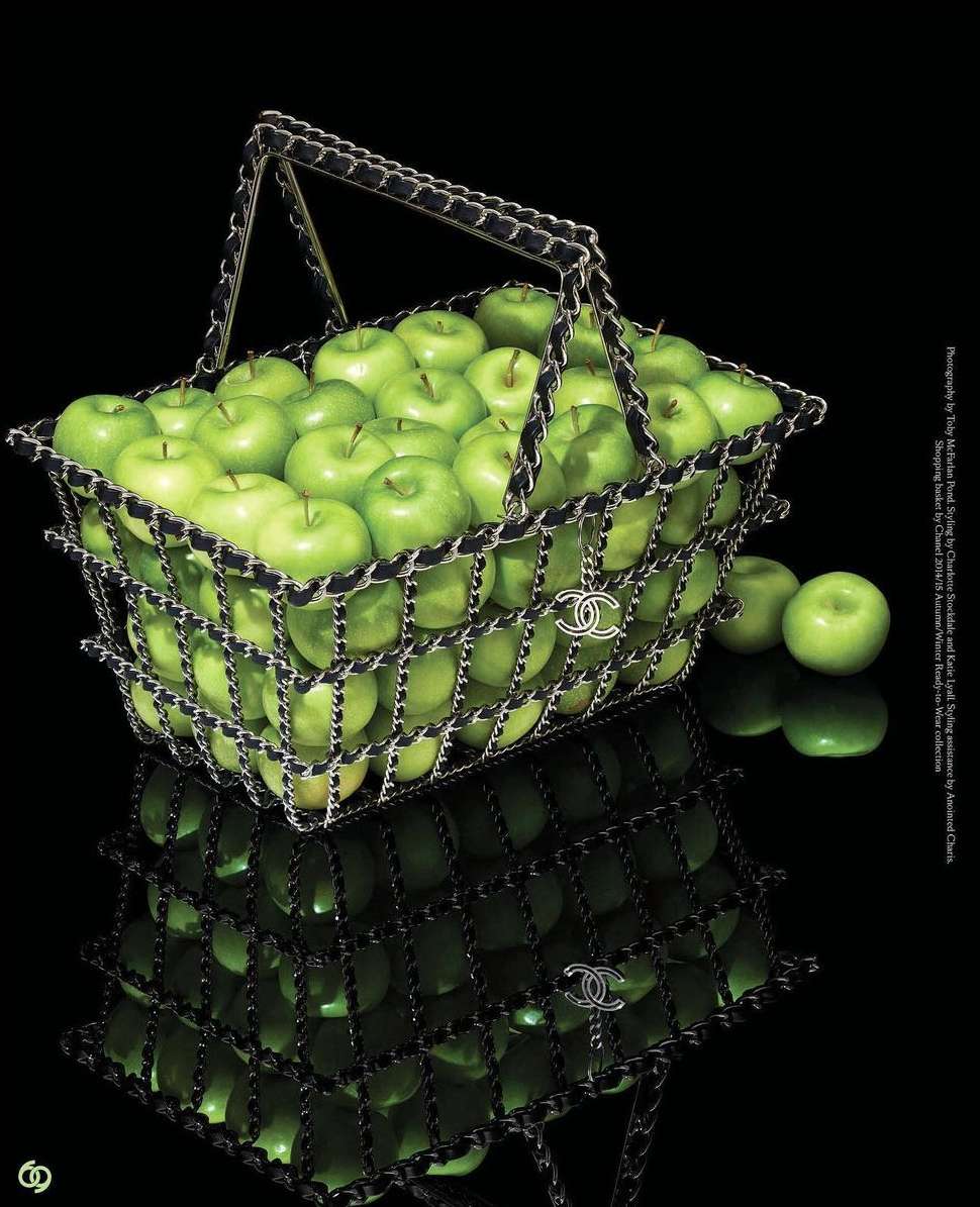 Overhead shot of a designer Chanel shopping basket filled with green apples, reflected on a black surface, luxury aesthetic.