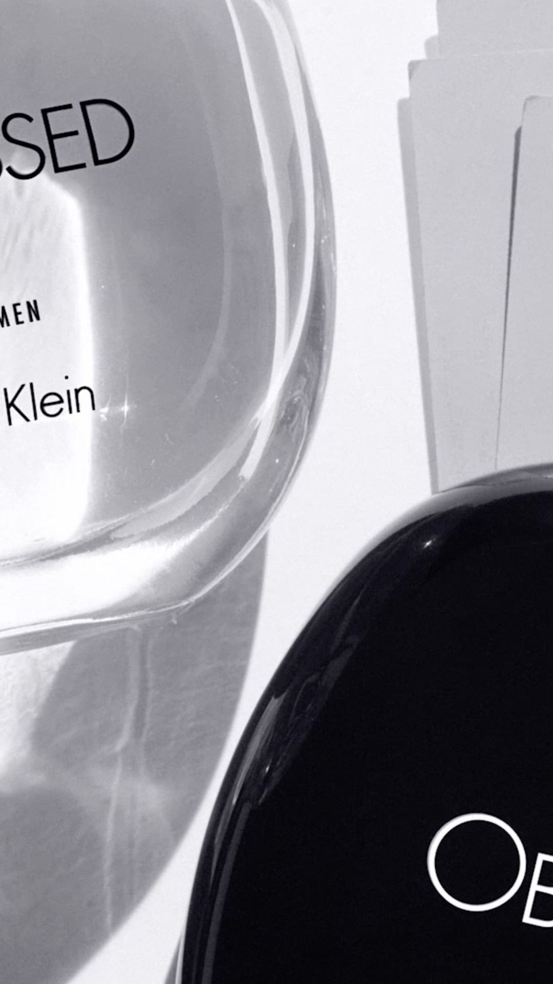 Close-up vertical shot of Calvin Klein OBSESSED perfume bottles in black and clear glass with white label text on a white background.