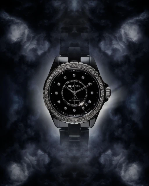 Elevated studio shot of a black Chanel J12 Automatic watch with a diamond bezel against a dark background with cloudy lighting.