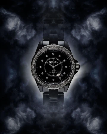 Elevated studio shot of a black Chanel J12 Automatic watch with a diamond bezel against a dark background with cloudy lighting.