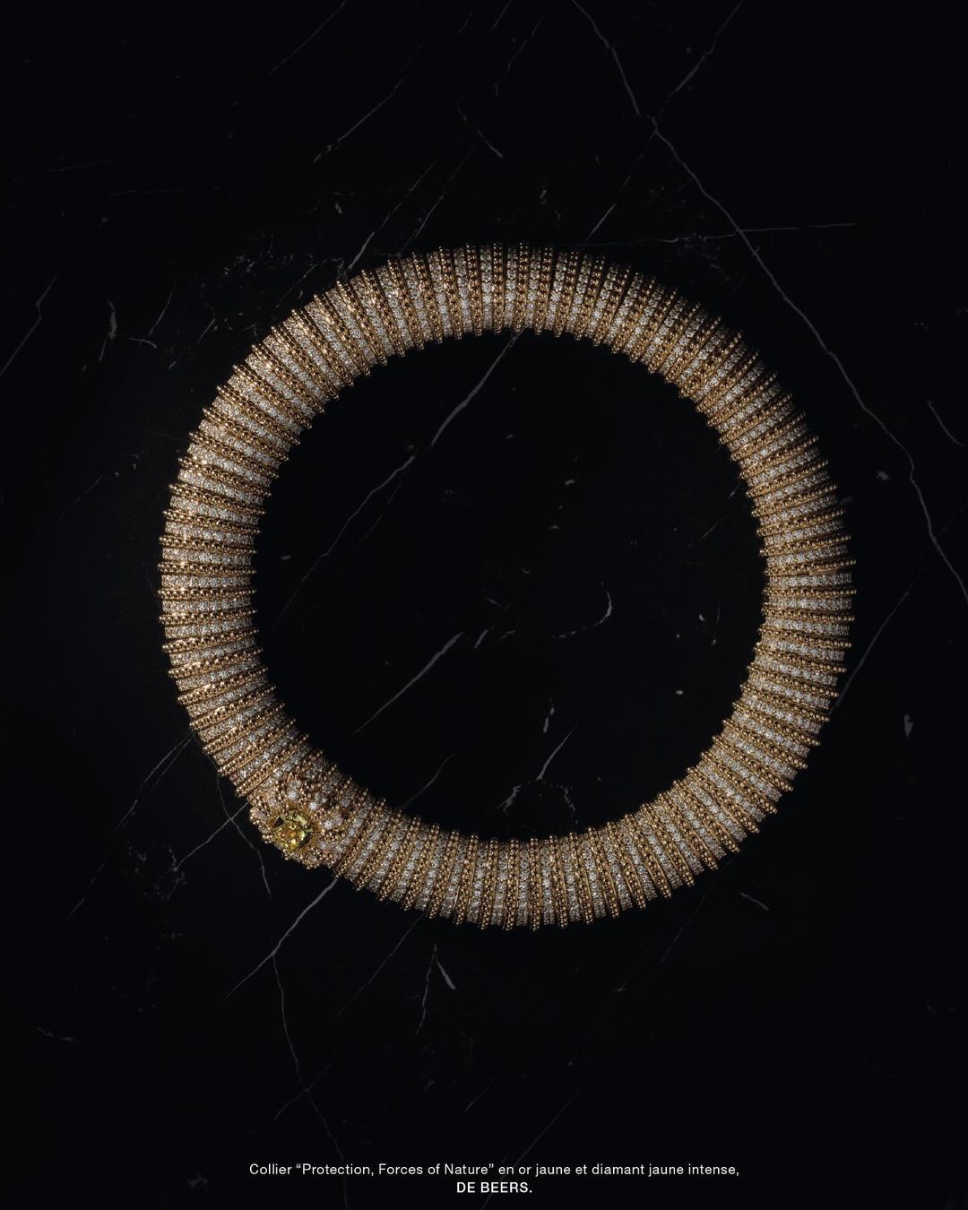 Close-up of a gold De Beers "Protection, Forces of Nature" collar necklace with diamond accents against a black marble background.