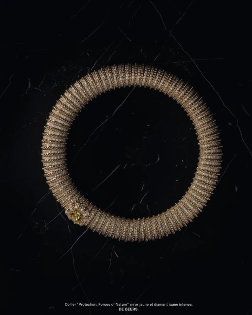 Close-up of a gold De Beers "Protection, Forces of Nature" collar necklace with diamond accents against a black marble background.