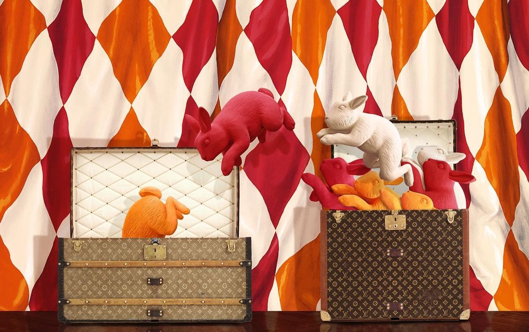 Two vintage Louis Vuitton trunks with colorful rabbit sculptures against a diamond-patterned backdrop, creating a whimsical and high-fashion still life.