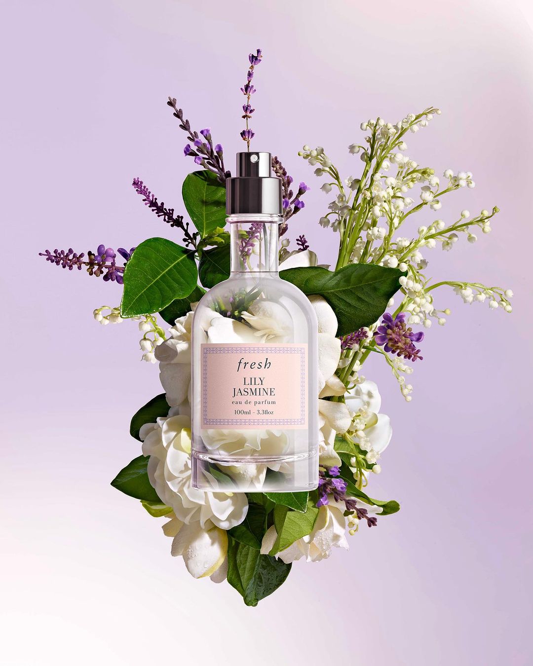 Elegant perfume bottle by Fresh with Lily Jasmine scent, surrounded by white and purple flowers on light purple backdrop. Product shot.