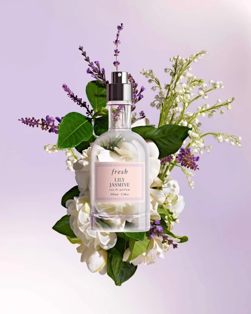 Elegant perfume bottle by Fresh with Lily Jasmine scent, surrounded by white and purple flowers on light purple backdrop. Product shot.