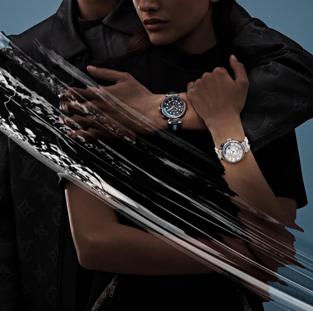 A couple poses in a studio setting with two Louis Vuitton watches. The man wears a black jacket with logo detail, the woman a black top.