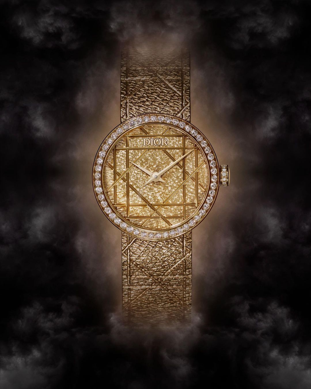 Elegant gold Dior watch with diamond bezel and textured band against a soft black smoky background in a beauty and fashion editorial shot.