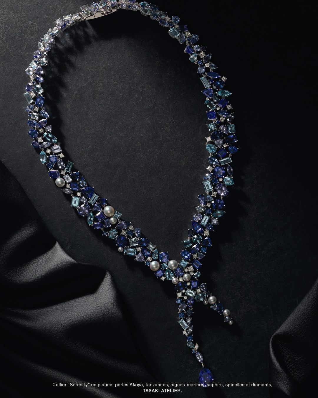 Elegant blue gemstone necklace made of tanzanite, sapphires, and pearls, displayed on a draped black fabric background in a jewelry advertisement.