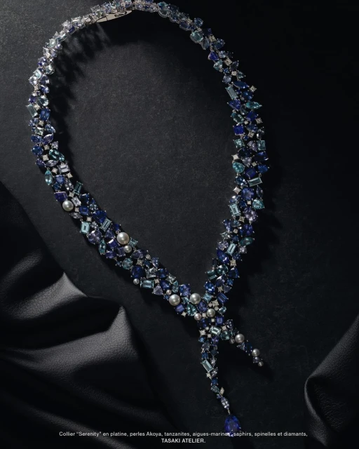 Elegant blue gemstone necklace made of tanzanite, sapphires, and pearls, displayed on a draped black fabric background in a jewelry advertisement.