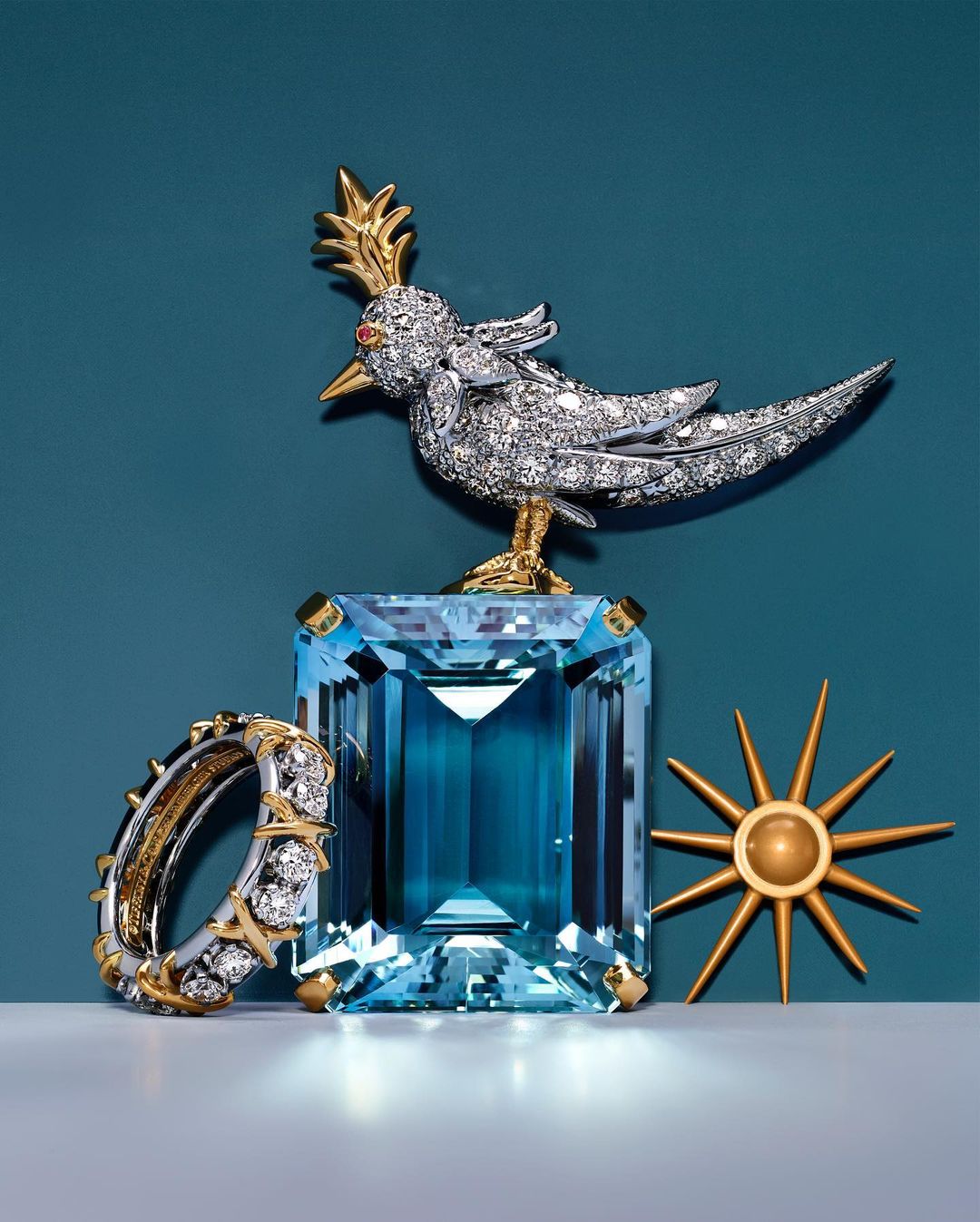 High-fashion jewelry still life shot with a diamond bird brooch atop a blue gemstone, complemented by a diamond ring and gold sunburst accent.
