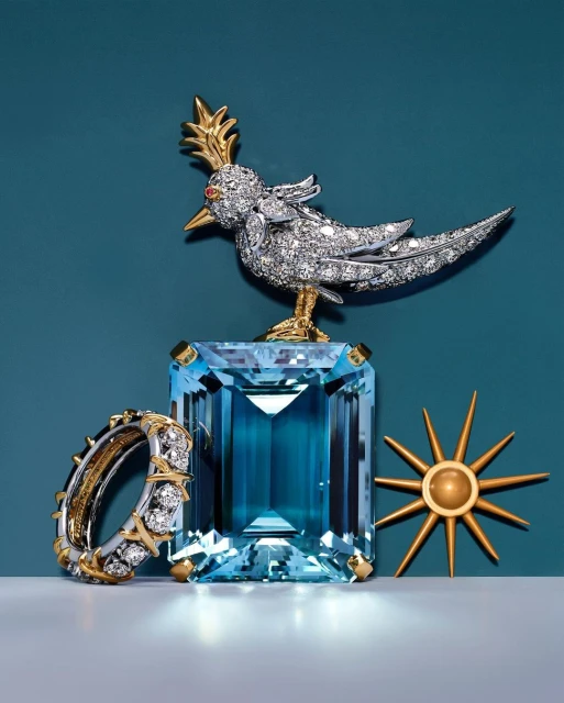 High-fashion jewelry still life shot with a diamond bird brooch atop a blue gemstone, complemented by a diamond ring and gold sunburst accent.