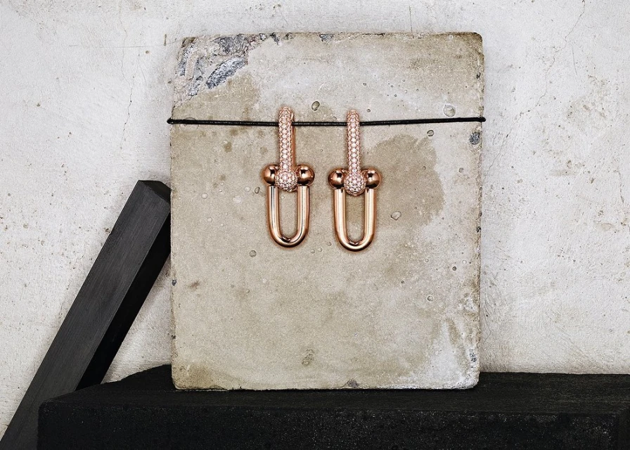 Studio shot of a pair of rose-gold-tone earrings with diamond accents displayed on a concrete surface with a dark background.