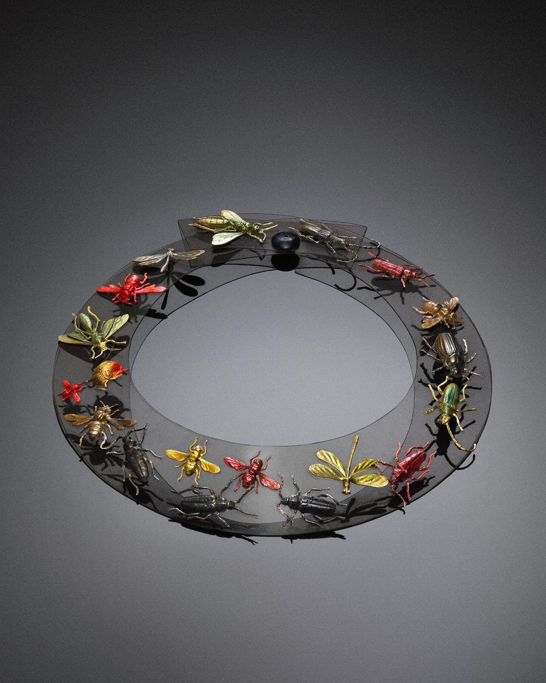 Black sheer collar necklace displaying colorful insect figures, shot in a photo studio against a grey background.