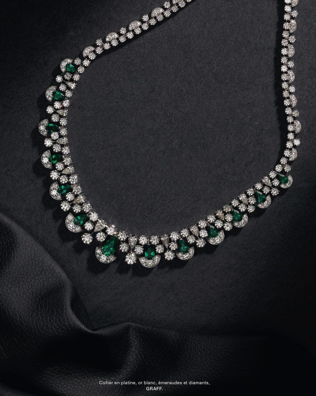 Elegant necklace featuring emeralds and diamonds against a draped black fabric background, showcased in a studio jewelry product shot.