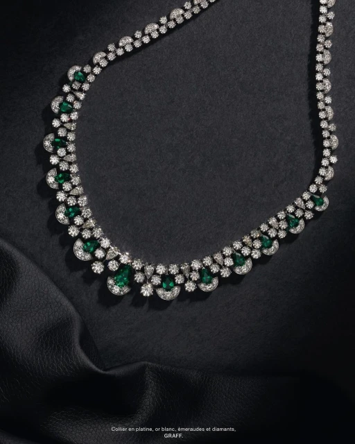 Elegant necklace featuring emeralds and diamonds against a draped black fabric background, showcased in a studio jewelry product shot.