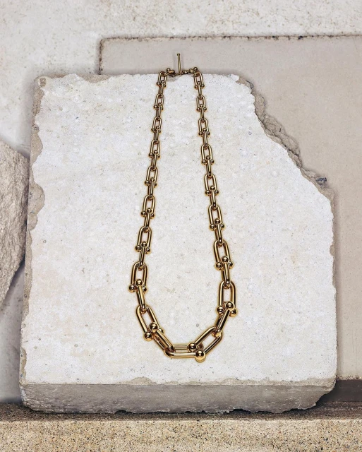 Close-up studio shot of a gold metal chain-link necklace, set against a textured white and beige stone background, fashion jewelry.