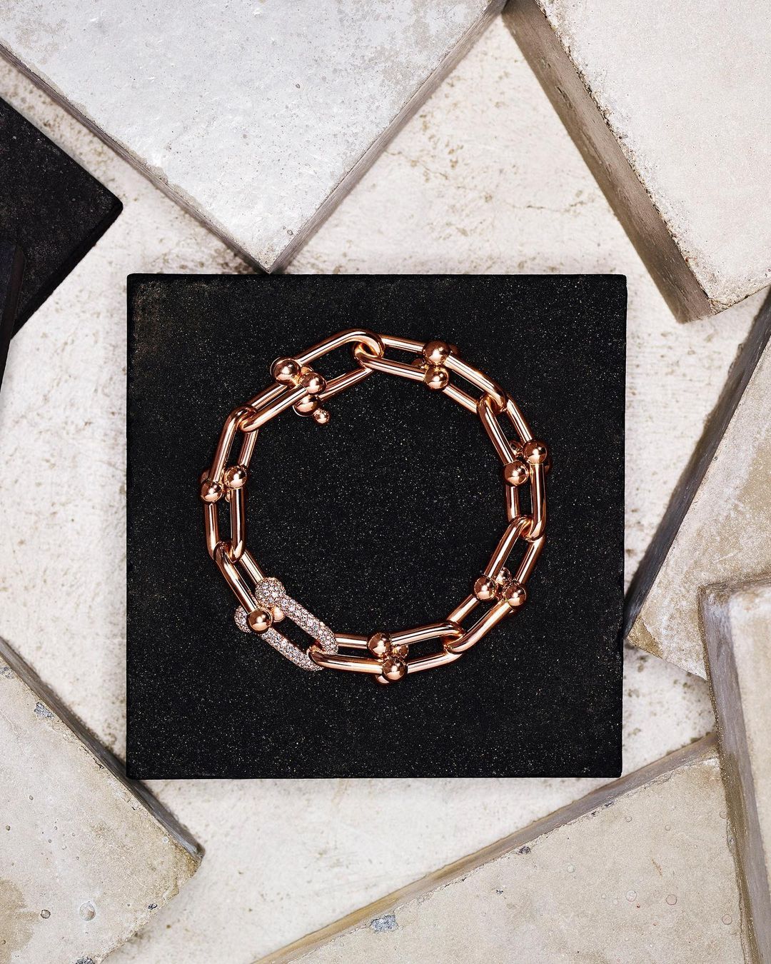 Close-up shot of a rose-gold chain-link bracelet with diamond clasp on a black surface, surrounded by textured stone tiles.