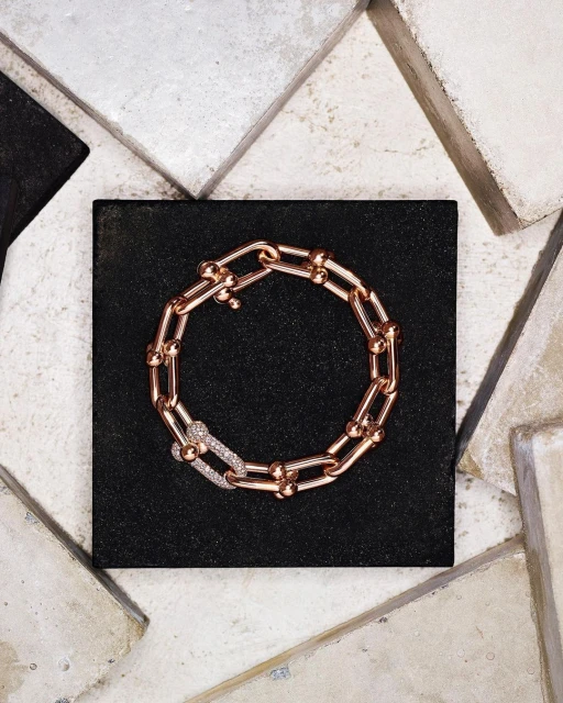 Close-up shot of a rose-gold chain-link bracelet with diamond clasp on a black surface, surrounded by textured stone tiles.