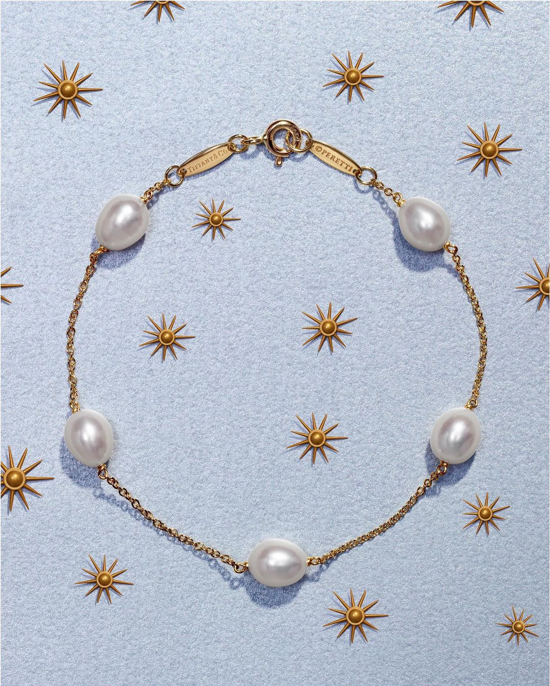 Gold chain bracelet featuring spaced pearls on a light blue background with gold starburst accents; fine jewelry fashion product shot.
