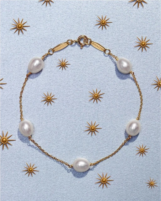 Gold chain bracelet featuring spaced pearls on a light blue background with gold starburst accents; fine jewelry fashion product shot.