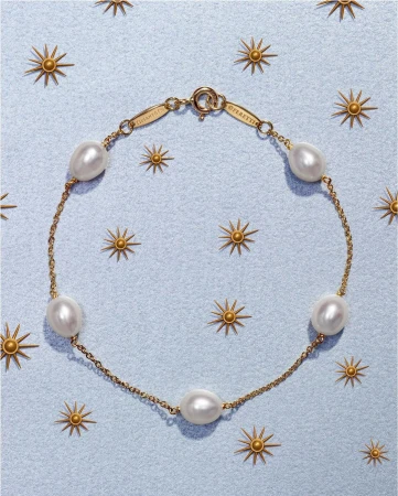 Gold chain bracelet featuring spaced pearls on a light blue background with gold starburst accents; fine jewelry fashion product shot.