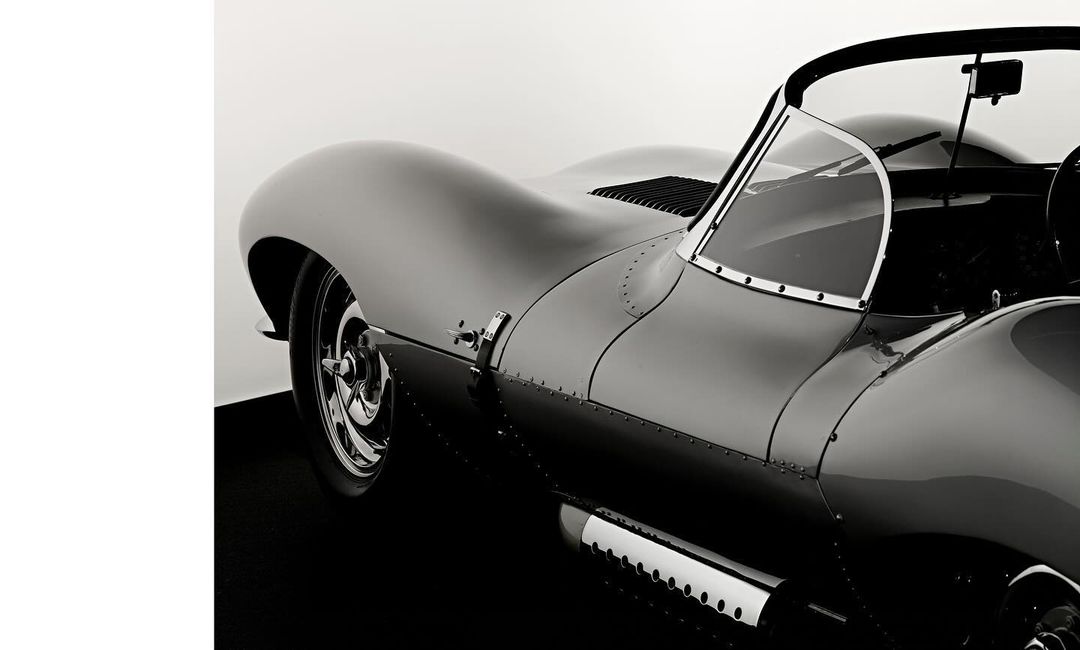 Black and white photograph of a Jaguar XKSS, a classic British sports car. The vehicle is parked on a black surface against a white backdrop.
