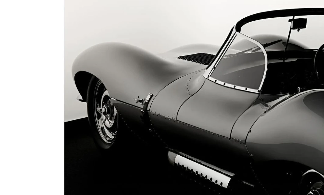 Black and white photograph of a Jaguar XKSS, a classic British sports car. The vehicle is parked on a black surface against a white backdrop.