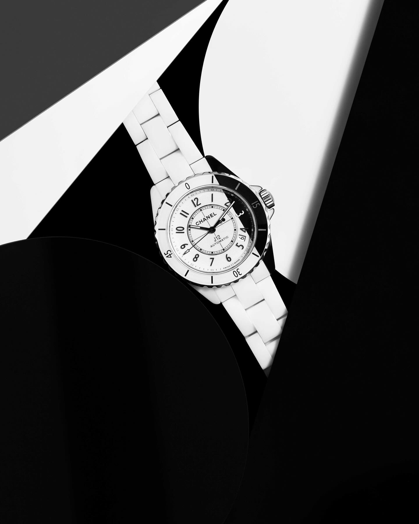 Close-up of a Chanel J12 watch with a white ceramic bracelet, set against a graphic black and white paper background creating contrast.