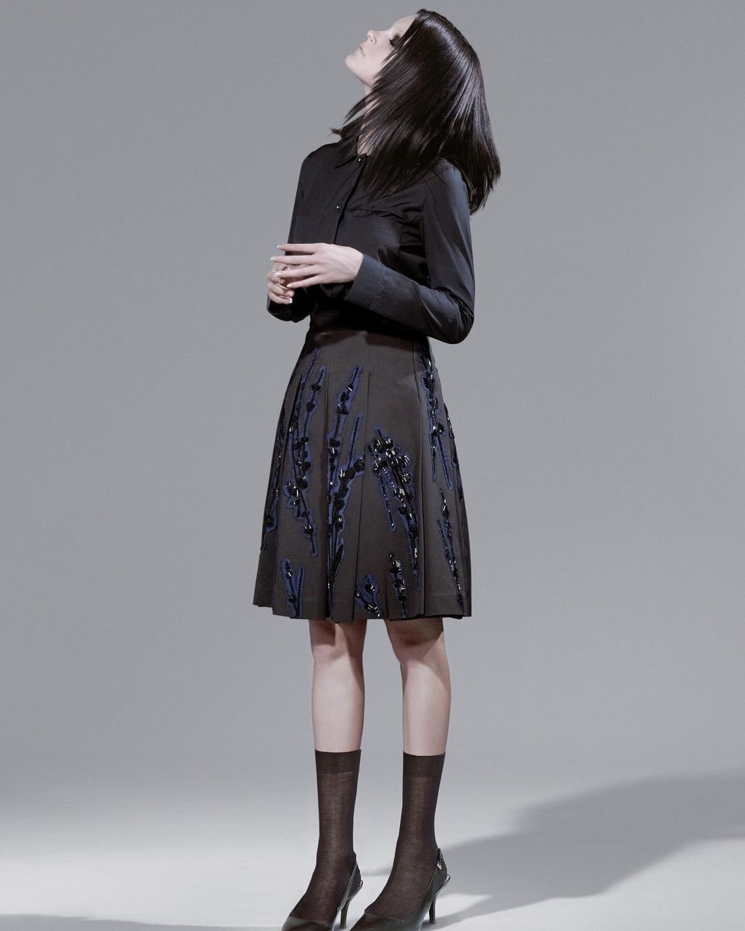 Elegant woman poses in black button-up shirt and A-line skirt with floral embellishments, styled with brown sheer socks and heels.
