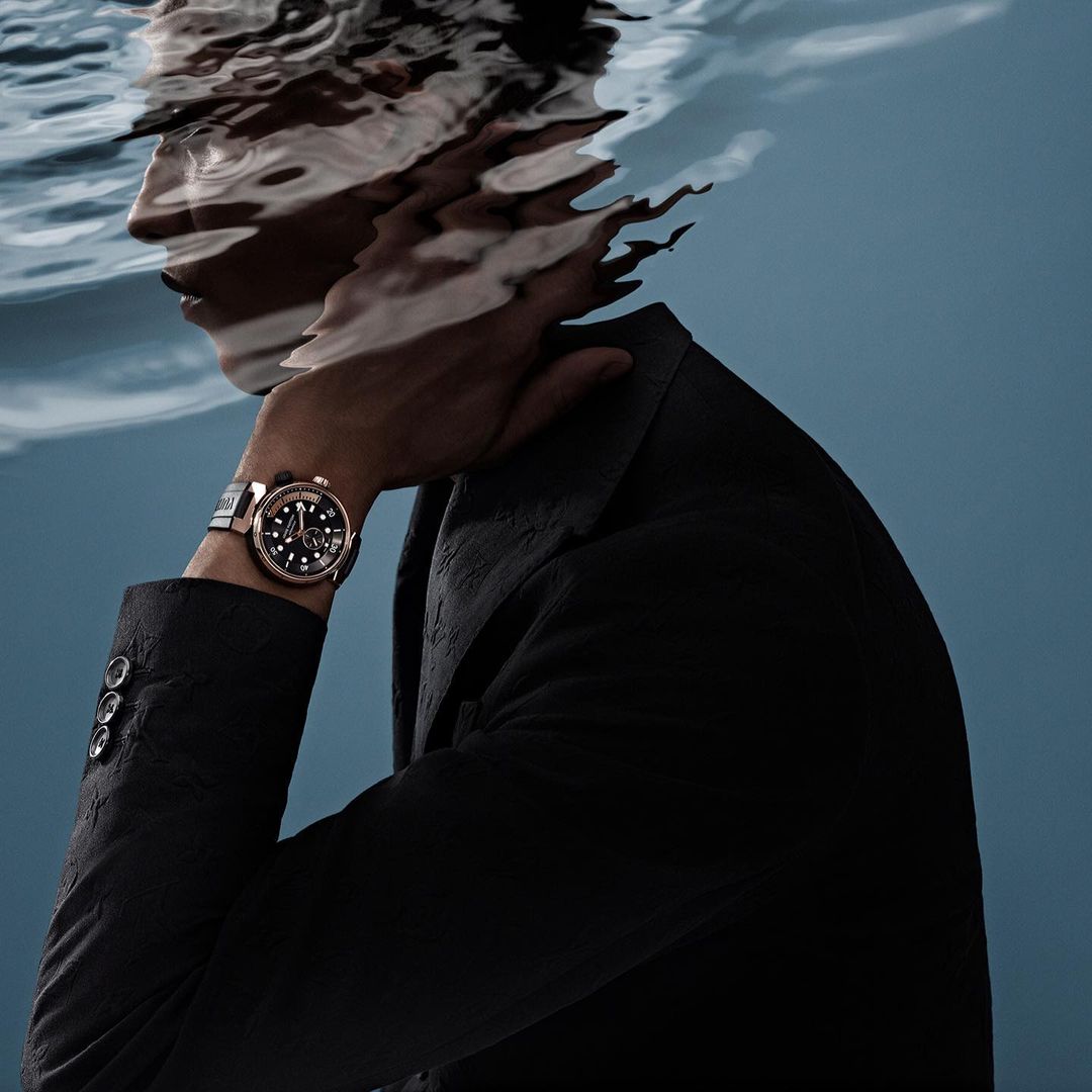 Model in suit wears a watch, partially submerged underwater with reflections in cool-tone studio lighting against a blue background.
