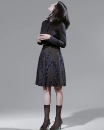 Model with dark hair standing in profile against a grey background wearing a black button-down shirt and a patterned midi skirt.