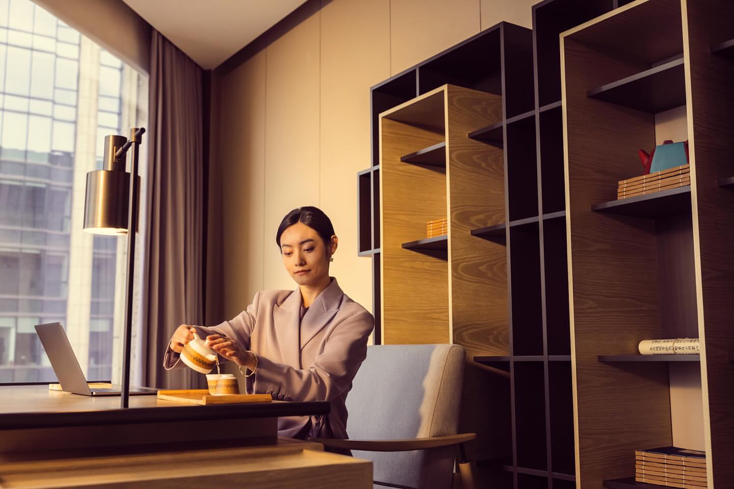 Young woman in blazer pours tea at modern desk with laptop, with contemporary bookshelf and city view through the window.