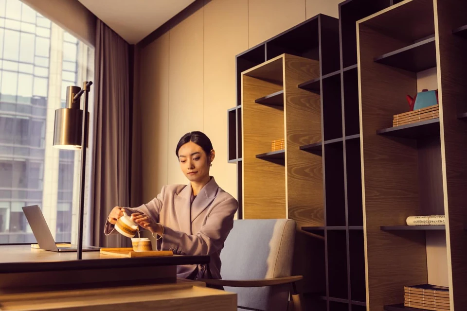 Young woman in blazer pours tea at modern desk with laptop, with contemporary bookshelf and city view through the window.